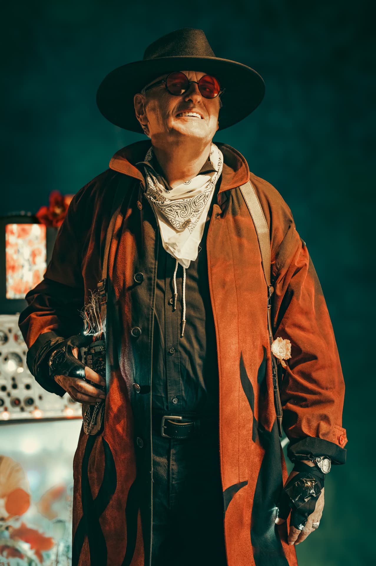 Chuck Wooke as The Traveler in a wide-brimmed hat, red round glasses, bandana and flame-painted rust coat, holding a wired circuit device before a dark teal backdrop