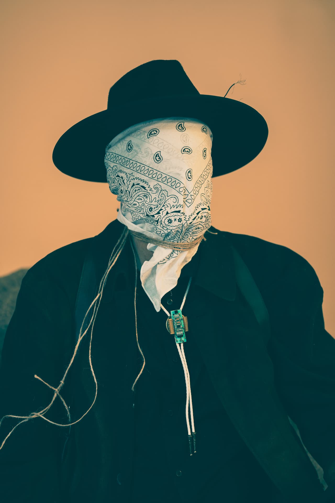Chuck Wooke as The Traveler, his face wrapped in a white paisley bandana tied with twine, wearing a black hat and a circuit-board bolo tie