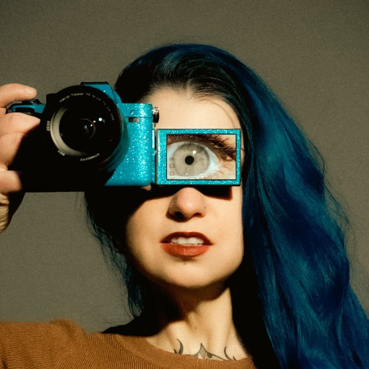 Katie Cooper with long blue hair holding a glitter-teal camera to her face, its flip screen showing her eye