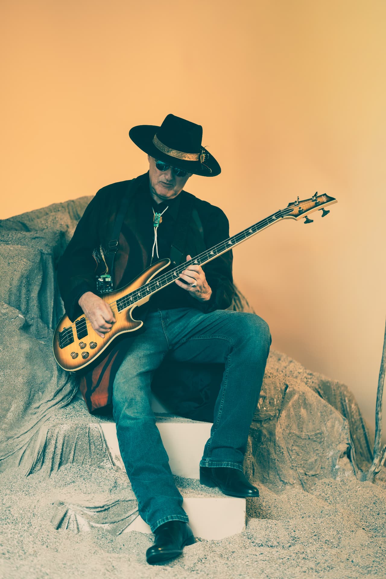 Chuck Wooke in black hat and sunglasses seated on sculpted desert rocks, playing a sunburst bass